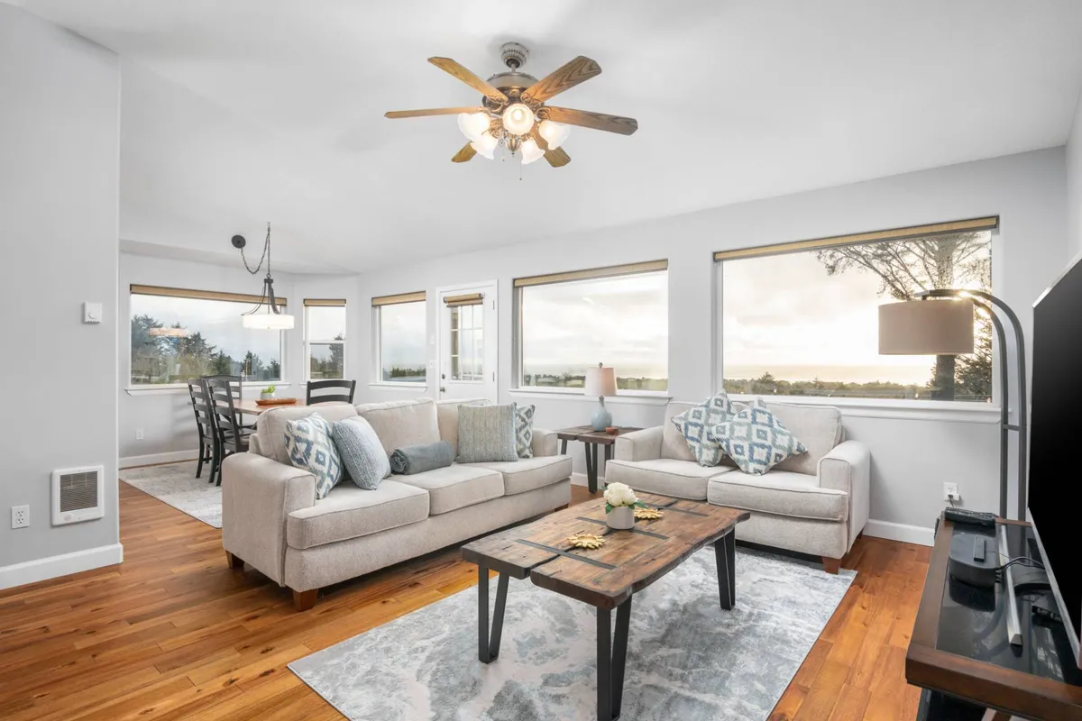 A warm, inviting living room featuring a plush sofa and coffee table set before expansive windows that frame stunning coastal views. The space is filled with natural light, accented by a ceiling fan above, creating the perfect spot for sunset watching, movie nights, and cozy gatherings.