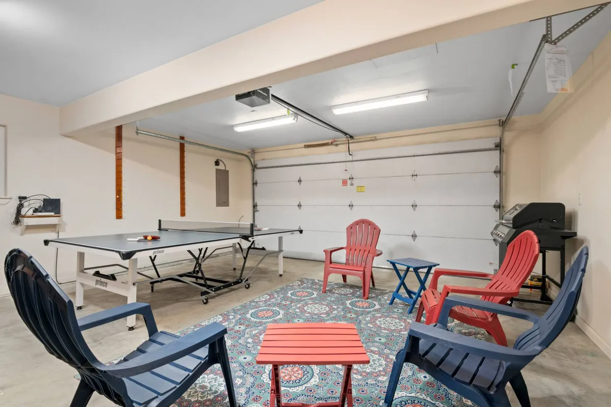This image captures the converted garage rec room, featuring a ping pong table at the center, colorful rug, Adirondack chairs for lounging, and a small red table. The space exudes a playful and relaxed atmosphere, ideal for friendly games and group activities.