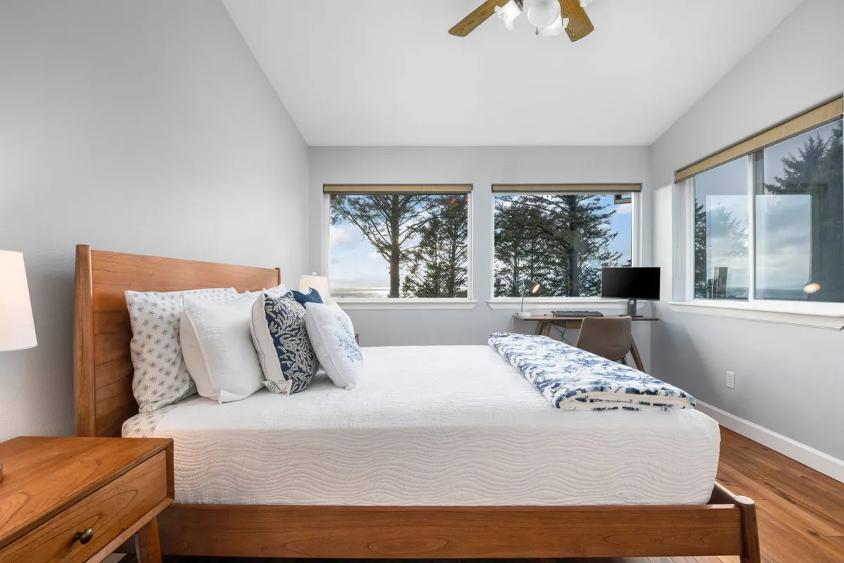 A bright bedroom with a bed featuring decorative pillows, large windows showcasing views of nature, and a functional desk area. The space is stylish yet practical, offering both relaxation and a spot for quiet work or reading.