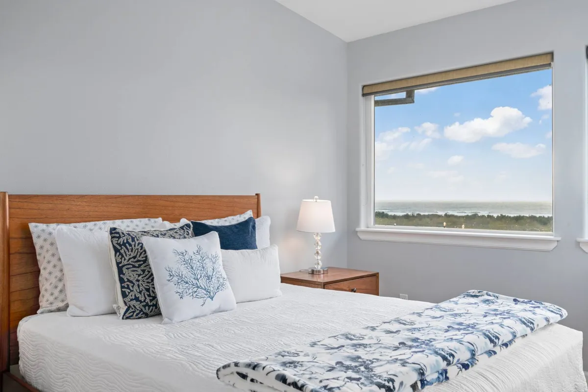 A serene bedroom featuring a bed adorned with decorative pillows, a window letting in natural light with a partial view outside, and a cozy nightstand with a lamp. The soft color palette and uncluttered styling reinforce a sense of restful retreat.