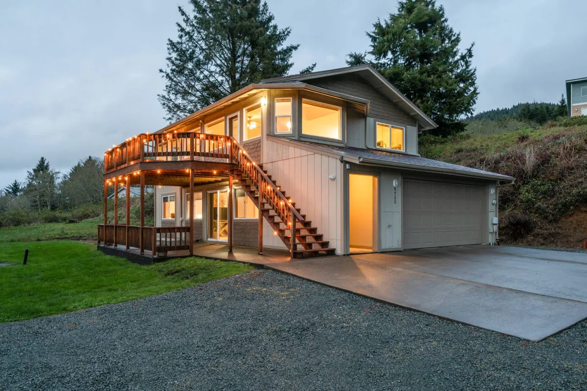 A welcoming exterior view showcasing a staircase with a railing leading up to a balcony adorned with string lights. Large windows suggest a bright, open interior, while the balcony offers an appealing spot to socialize and survey the surrounding coastal environment.