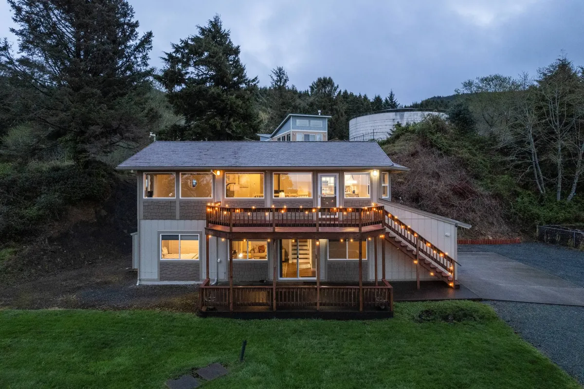 An exterior evening view of Casa Del Sol features a welcoming wooden deck lit by warm string lights. Large windows reveal cozy indoor spaces, while the deck itself is surrounded by trees, inviting guests to gather, relax, or stargaze under the night sky. The ambiance is both cozy and invigorating, suggesting the property’s potential for both quiet reflection and social connection after work hours.