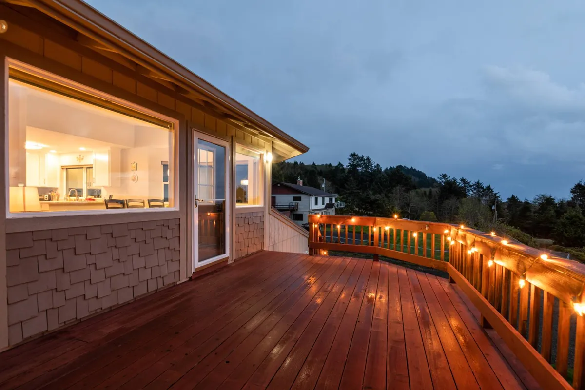 This image features the spacious wooden deck of Casa Del Sol, illuminated by string lights, with large windows providing a glimpse into the inviting interior. The setting is perfect for outdoor gatherings, watching sunsets, or sharing a morning coffee while overlooking the ocean.