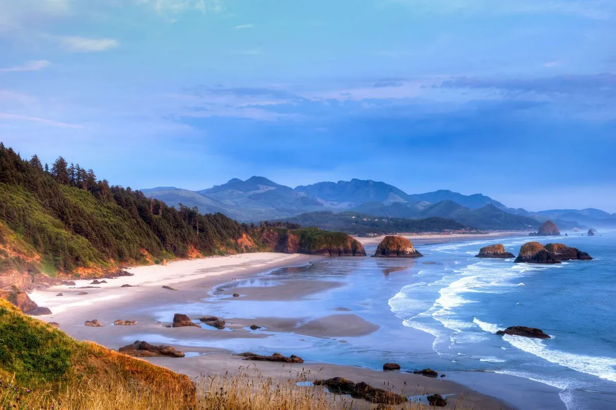 A sweeping view of a sandy beach edged by dramatic rock formations, rolling ocean waves, and a distant forested mountain range. The scene captures the essence of Oregon’s rugged coastline and highlights the natural playground available for families, with open sands perfect for beach games and exploration.