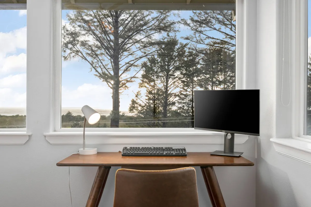 Inside, a wooden desk faces large windows with views of lush trees outside. The setup includes a computer monitor and lamp, creating a comfortable, productive workspace flooded with natural light. The serene backdrop and practical furnishings highlight the space’s suitability for focused work while staying connected to the surrounding nature.
