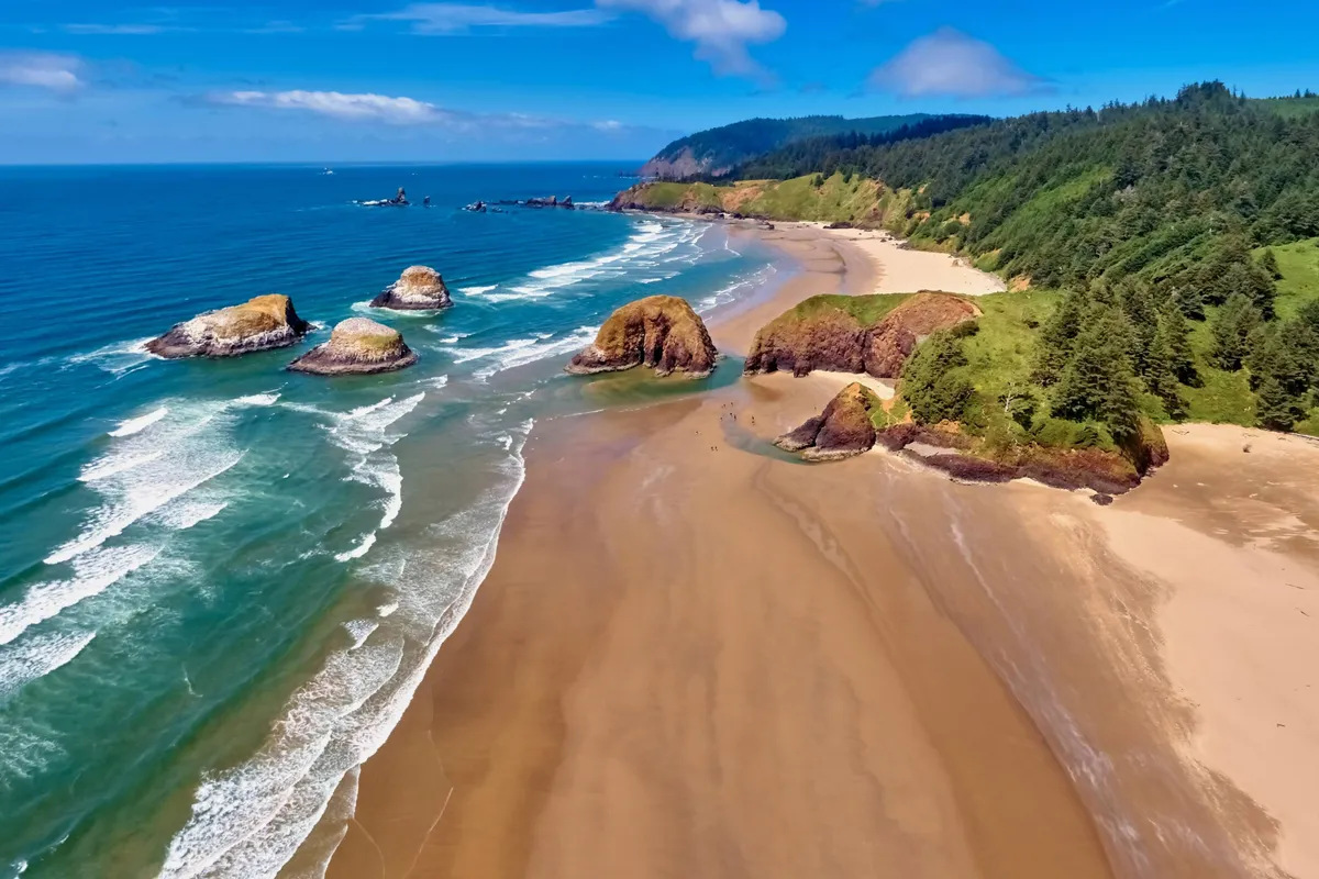 A picturesque beach scene with sunlit rock formations, gentle waves, green hills in the background, and smooth sandy stretches. The foreground offers ample room for building sandcastles, running, or simply enjoying the serene oceanfront vibe.